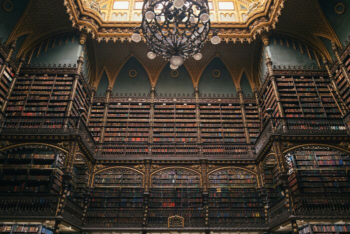 Ornate grandiose library interior with towering bookshelves and intricate gold detailing under a decorative chandelier.
