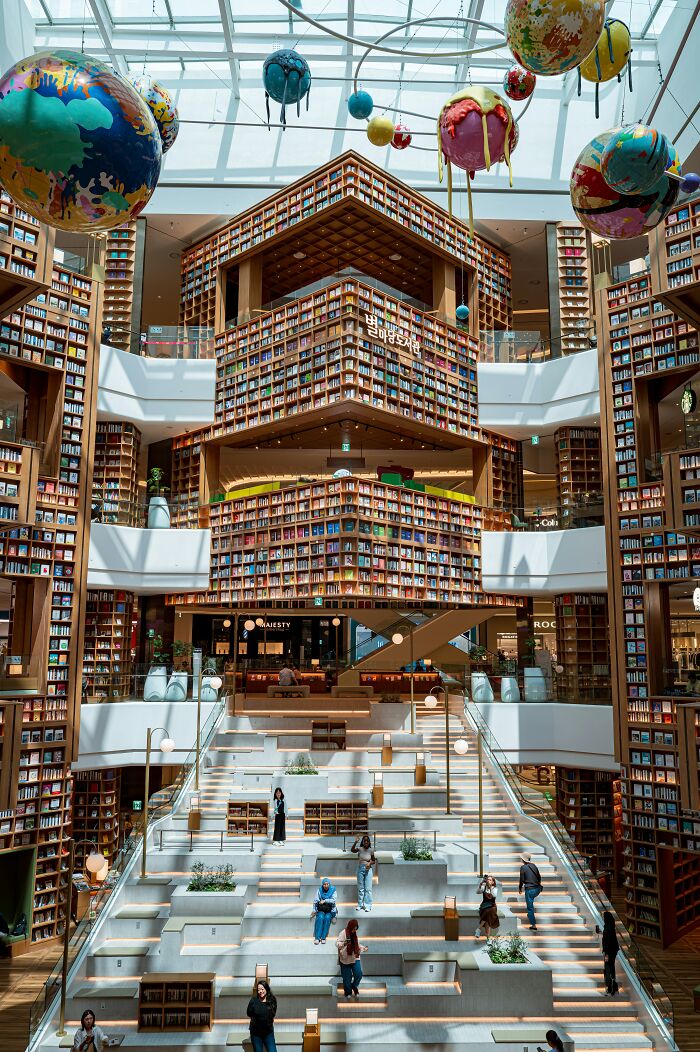 Grandiose library interior with towering bookshelves, large skylight, and colorful planet sculptures hanging from ceiling.